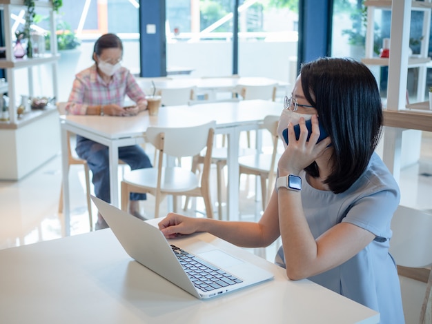 Photo two asian women wearing face mask and using smartphone and laptop for video calling or working, sit on separate tables for keeping safety social distancing, as new normal lifestyle concept.