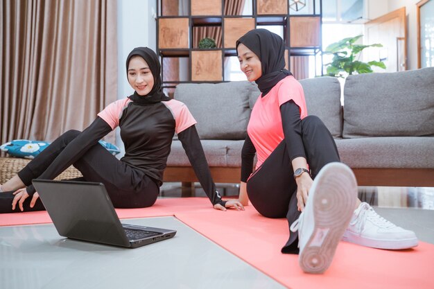 Two asian women wearing hijab sportswear sit on the floor while warming up their hips together in the house