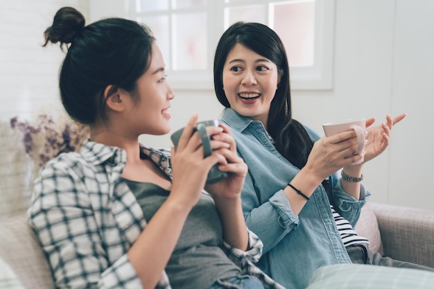 Two asian young girls roommates relaxing on sofa at home
drinking hot tea looking outside enjoy the city view from window on
sunny day. women stay indoors lazy chatting talking on couch on
weekends.