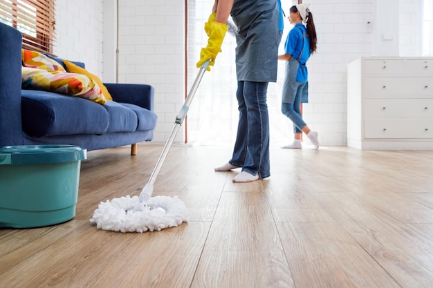 Two Asian young professional cleaning service women worker team working in the house The girl housekeeper wiped a wet mop on wooden floor with another one clean the window with a feather duster