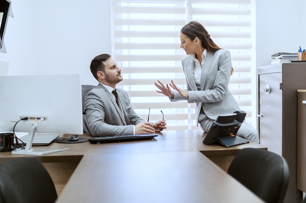 Two attractive Caucasian colleagues dressed in formal wear sitting in office and chatting.