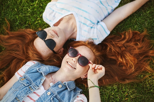 Two attractive european girlfriends with red natural hair and shiny smile grinning from happiness while lying on grass and gazing in sunglasses and clouds. Lifestyle and people concept