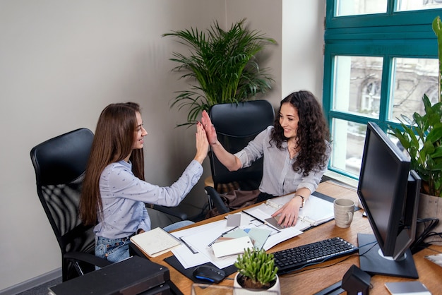 Two attractive female designers in blue shirts working together with new project on pc in the modern office. The girls are clapping one another in the palm.