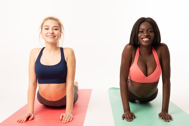 Foto due ragazze africane ed europee focalizzate in forma attraente vestite in abiti sportivi in piedi in posizione yoga cobra bassa