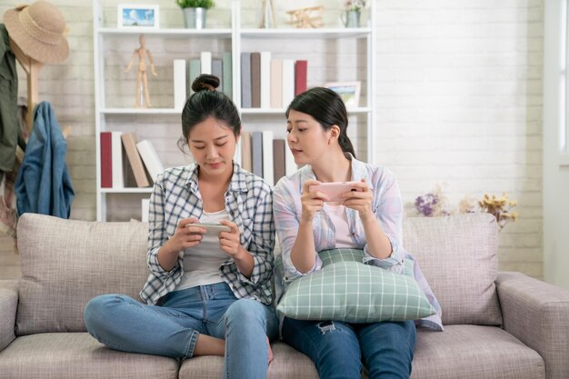 スマートフォンでゲームをしているソファに座っている2人の魅力的なガールフレンド。不思議なことに彼女の姉妹の携帯電話の画面を覗く若い女性。ソファの家でインターネット上で楽しい競争をしている女性のグループ