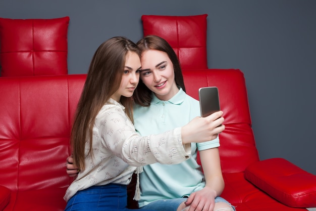 Two attractive girlfriends sitting on red leather couch and makes selfie on phone camera