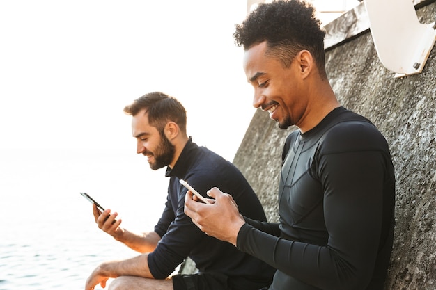 Two attractive smiling young healthy sportsmen outdoors at the beach, using mobile phones