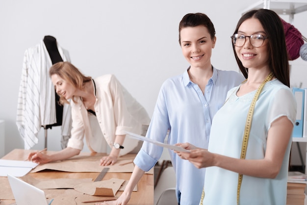 Two attractive stylish women standing near tble in the atelier while their colleague making notes on patterns