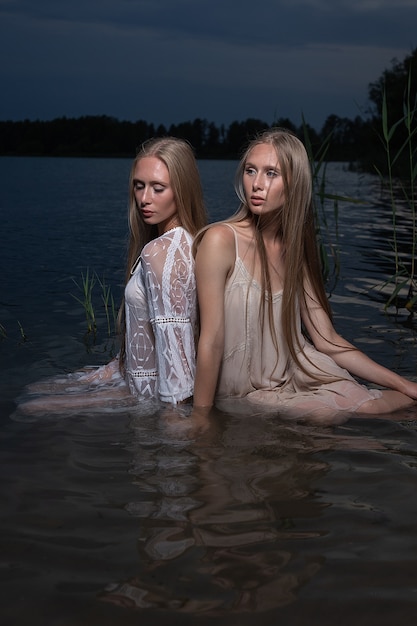 Photo two attractive young blond twin sisters posing in light dresses in water of lake at summer night.