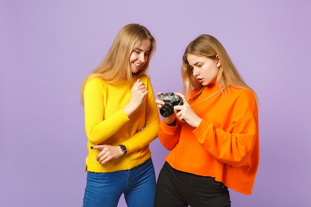 Two attractive young blonde twins sisters girls in vivid colorful clothes holding retro vintage photo camera isolated on violet blue wall. People family lifestyle concept. 