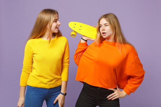 Two attractive young blonde twins sisters girls in vivid colorful clothes holding yellow skateboard isolated on pastel violet blue wall. People family lifestyle concept.