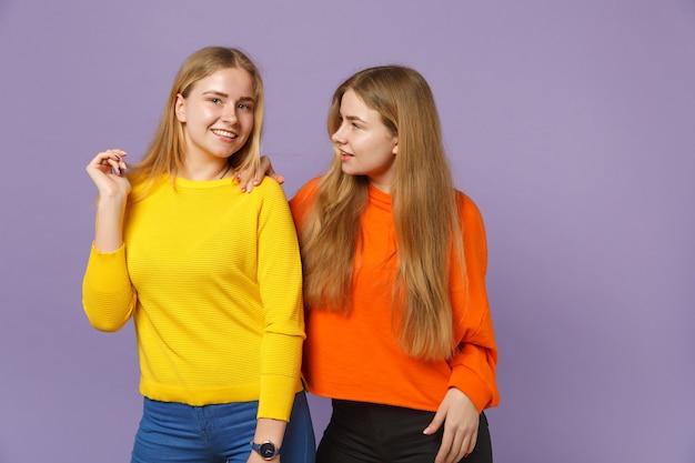 Two attractive young blonde twins sisters girls in vivid colorful clothes standing,  isolated on pastel violet blue wall . People family lifestyle concept. 