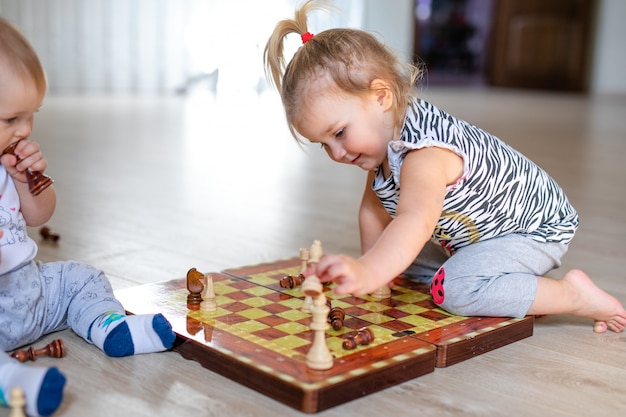 Two babies boy and girl playing chess on the white wooden floor at home.