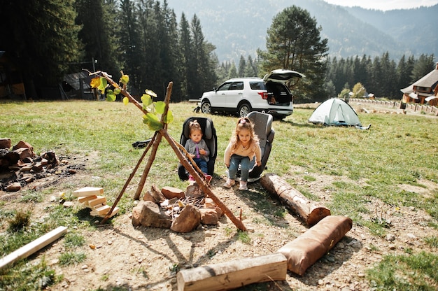 Two baby girl sitting by the fire in mountain. autumn hike\
weather. warming and cooking near flame together.