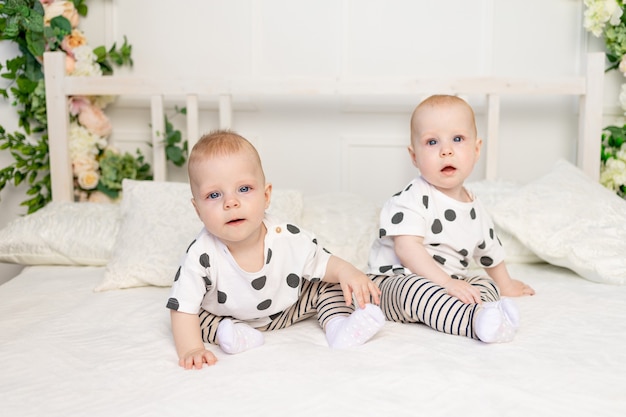 Two baby twins sitting on the bed in the same clothes, brother-sister relationship, fashionable clothes for children of twins, concept of marriage and friendship