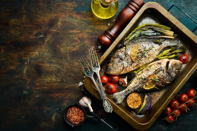 Two baked dorado fish with asparagus and vegetables in a metal tray Top view Free copy space On a black stone background