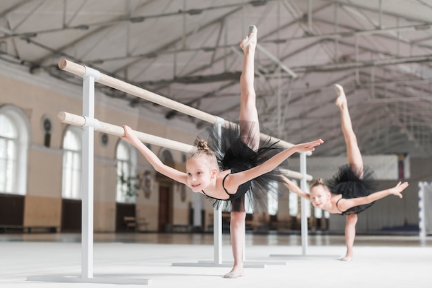 Two ballerina girls stretching their legs up with barre support in dance class