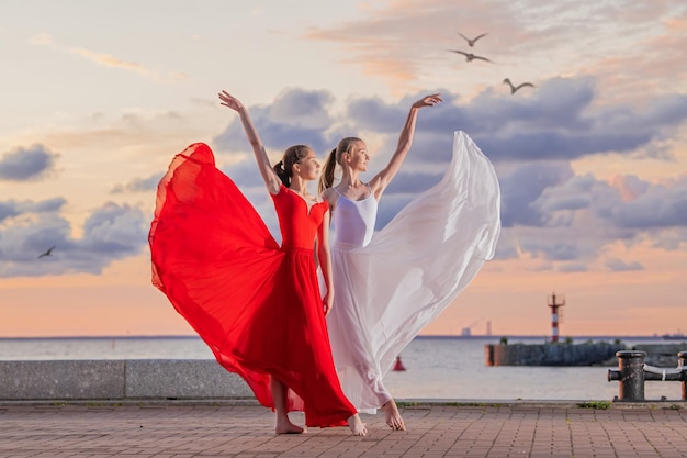 Two ballerinas in a white and red flying skirt and leotard dancing in a duet on the embankment of the ocean or sea against the backdrop of the sunset sky