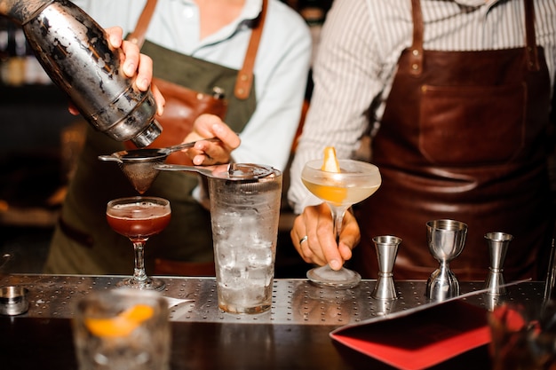 Two barmen in aprons at the bar counter prepare alcoholic cocktail with ice , using shaker and strainer