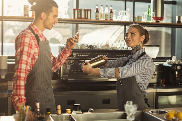 Two bartenders recording tutorial video on smartphone at coffee shop. Young barman shooting professional tattoed barista while she making cocktail. Skill share, technology and small business concept