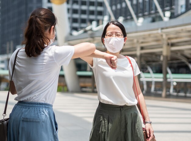 two beautiful asian women greeting each other with elbow, wearing a disposable medical face mask