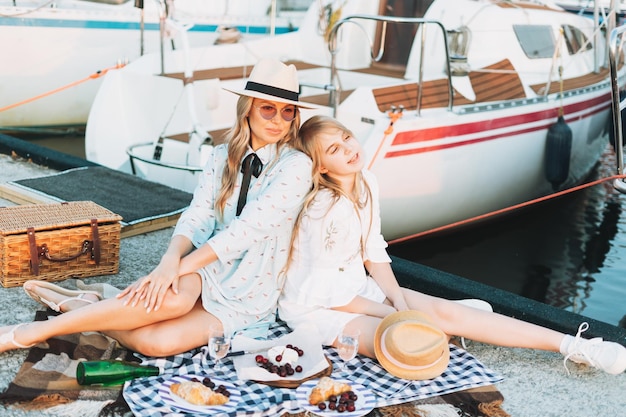 Two beautiful blonde girls friends mother and daughter in straw hats having picnic on the yacht pier