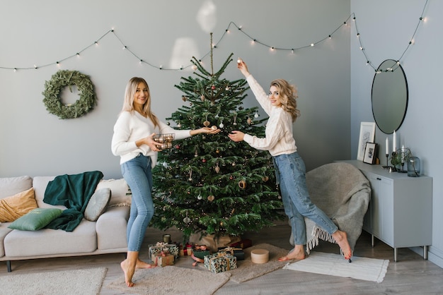 두 명의 아름다운 금발 자매가 크리스마스 트리를 장식하는 축하 준비를 하며 즐거운 시간을 보내고 있습니다.