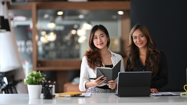 Two beautiful businesswoman sitting together in modern office and smiling to camera