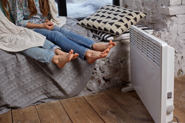 two beautiful caucasian little sisters sitting on the windowsill, covered by blanket. they froze up and trying to warm up, using heater near their foots