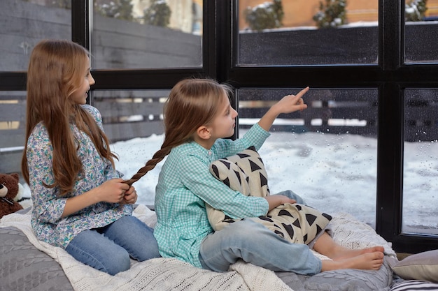 Two beautiful caucasian sisters sitting on the windowsill, they saw something interesting at the window and try to see it