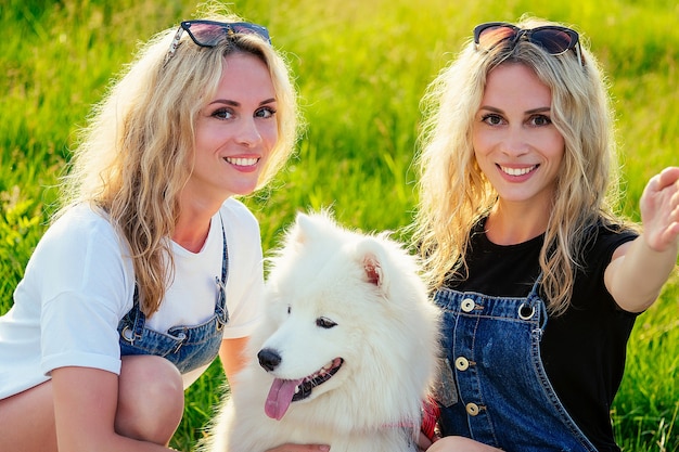 公園で電話で座って自分撮りをしている白いふわふわのサモエド犬と2人の美しく魅力的な金髪の双子の女性。