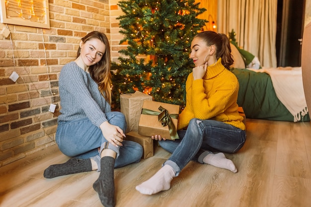 2人の美しい陽気な幸せな若い女の子のガールフレンドが自宅で新年の木の背景にクリスマスプレゼントを贈る