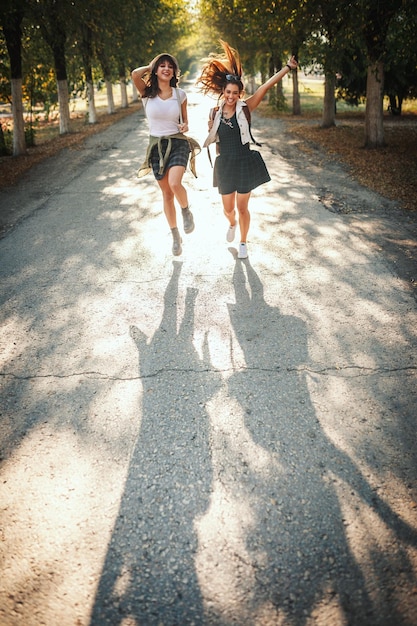 Foto due belle giovani donne allegre con gli zaini sulla schiena stanno camminando lungo il viale soleggiato autunnale e si tengono per mano.