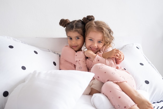 Две красивые разнообразные дети девочки в пижамах обнимаются на кровати в современной светлой квартире.
