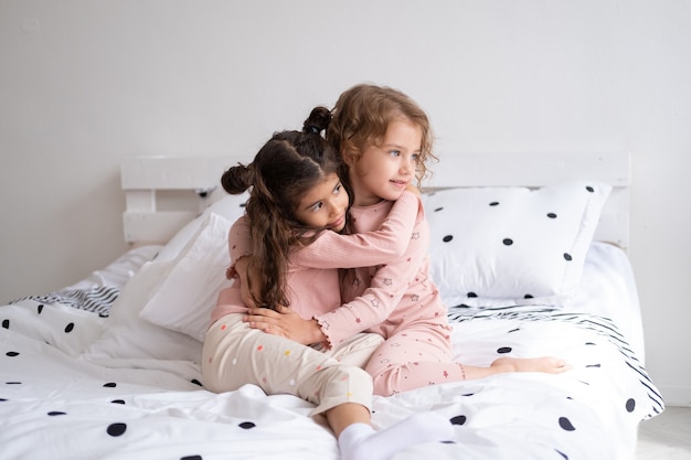 Two beautiful diverse kids girls in pajamas hugging on bed in modern bright apartment.