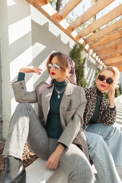 양복 스웨터 청바지와 표범 재킷으로 트렌디한 아웃웨어를 입은 두 명의 아름다운 패션 걸스 힙스터가 거리 도시 가을 여성 스타일의 옷을 입고 앉아 있다