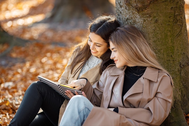 Two beautiful female friends spending time together. Two young smiling sisters talking and standing near tree with a book. Brunette and blonde girls wearing coats.