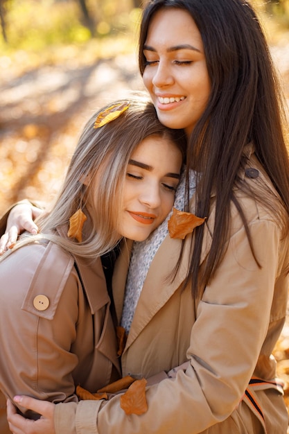 Two beautiful female friends spending time together. Two young smiling sisters walking in autumn park and hugging. Brunette and blonde girls wearing coats.