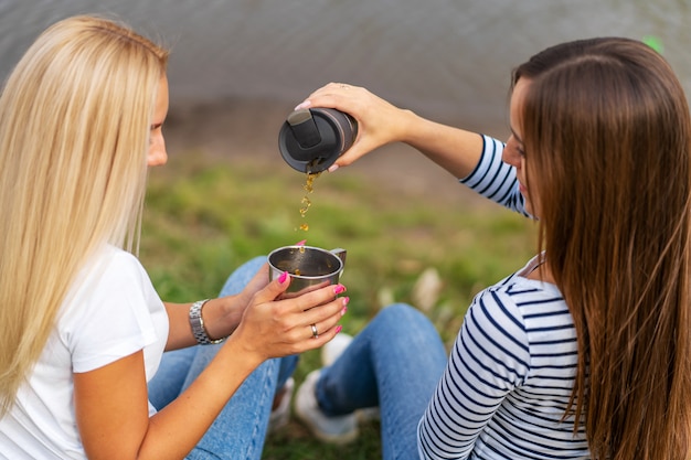 Two beautiful girls enjoy nature and drink hot tea on lakeshore with beautiful view