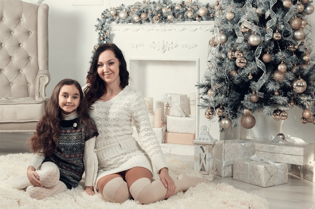 Two beautiful girls, mother and daughter siting on a floor in Christmas decorated room.