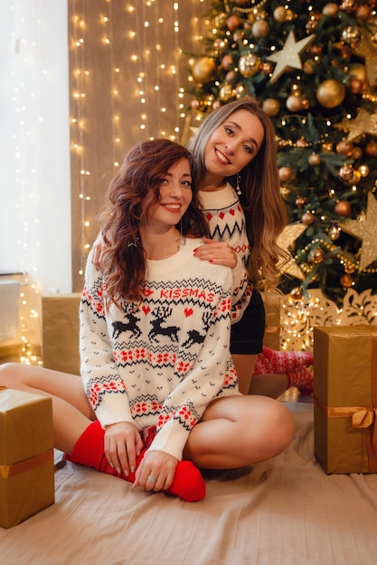 Due belle ragazze alla vigilia di capodanno. migliori amiche della giovane donna che celebrano il natale in casa. l'amicizia di divertimento non finisce mai il concetto. bellissime decorazioni natalizie dorate su un alto albero di natale