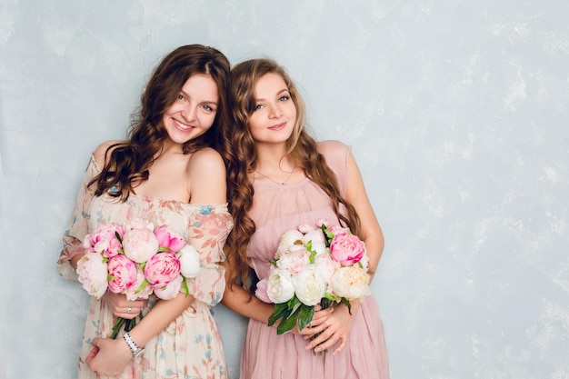 Two beautiful girls stand in a studio and hold bouquets of flowers. They wear light silk dresses. One is blond, and one is brunette. Both have curly hair.