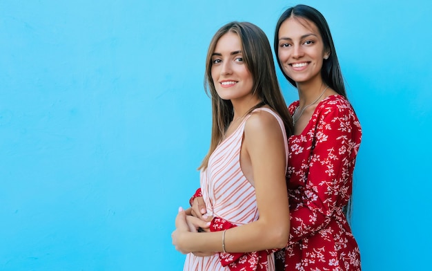 Two beautiful girls in summer dresses are hugging and holding hands while standing in profile, smiling and looking in the camera