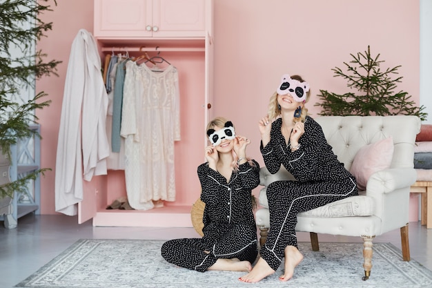 Foto due belle ragazze bionde, felici e sorridenti, in pigiama alla moda e in eleganti maschere da notte a forma di panda, si siedono sul divano vintage e sul pavimento e in posa all'interno