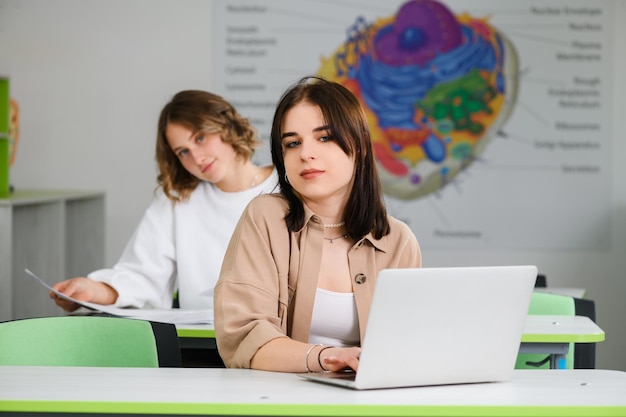 Two beautiful high school female teenage friends sitting on desks in modern class with laptop and notebook and look at camera