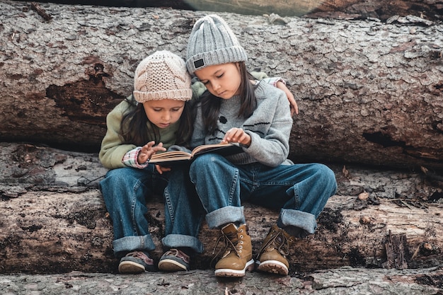 Two beautiful little girls reading books in the autumn forest , sitting on a log. The concept of education and friendship.