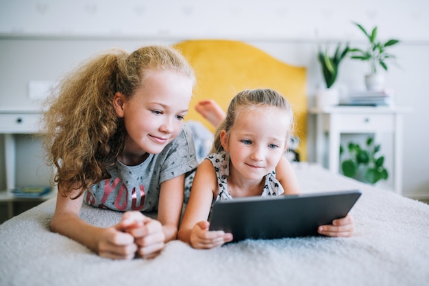 Two beautiful little sisters lying in the bed and look at the screen of a tablet, smart kids using smart technology