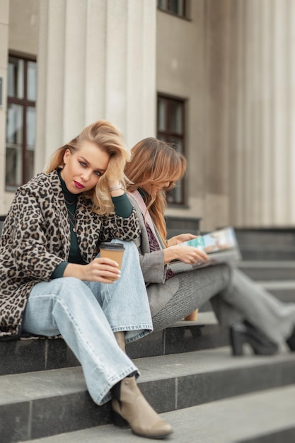 Две красивые девушки-модели в модной одежде сидят на ступеньках в городе и отдыхают с кофе и журналом