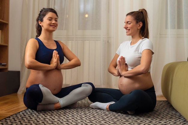 Two beautiful pregnant woman mediating and keeping eyes closed while sitting in lotus position