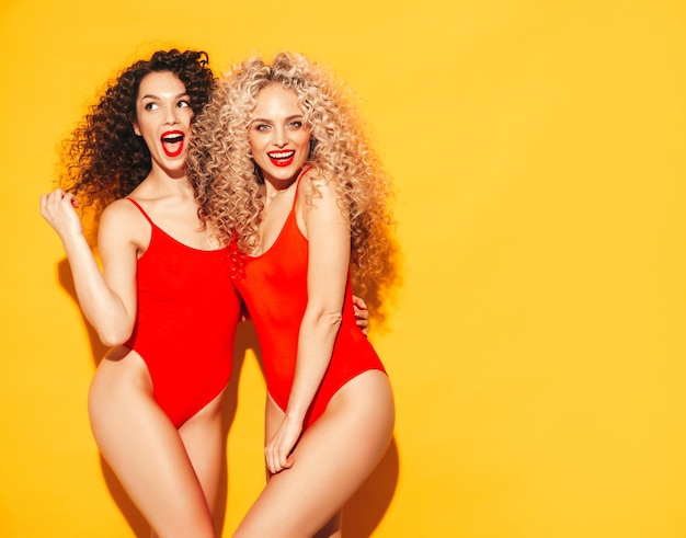 Two beautiful sexy smiling hipster women in red summer swimwear bathing suits Trendy models with afro curls hairstyle having fun in studio Hot female isolated on yellow in sunglasses